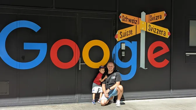 Me crouching with my son in front of a large Google logo on a black wall at the Google Zurich office, beside a signpost reading Schweiz, Suisse, Svizzera and Svizra